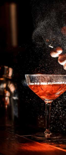 Bartender in black apron and blue shirt sprays an orange peel in cocktail glass with ice at a bar counter