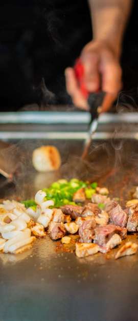chef's hands with spatula over teppanyaki. cooking vegetables meat and seafood on hot hibachi grill table. Traditional Japanese Cuisine. Teppan show